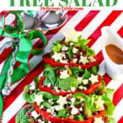 A Christmas tree salad on a red and white striped table with a white pitcher filled with cranberry vinaigrette and serving tongs tied with a red and green bow.