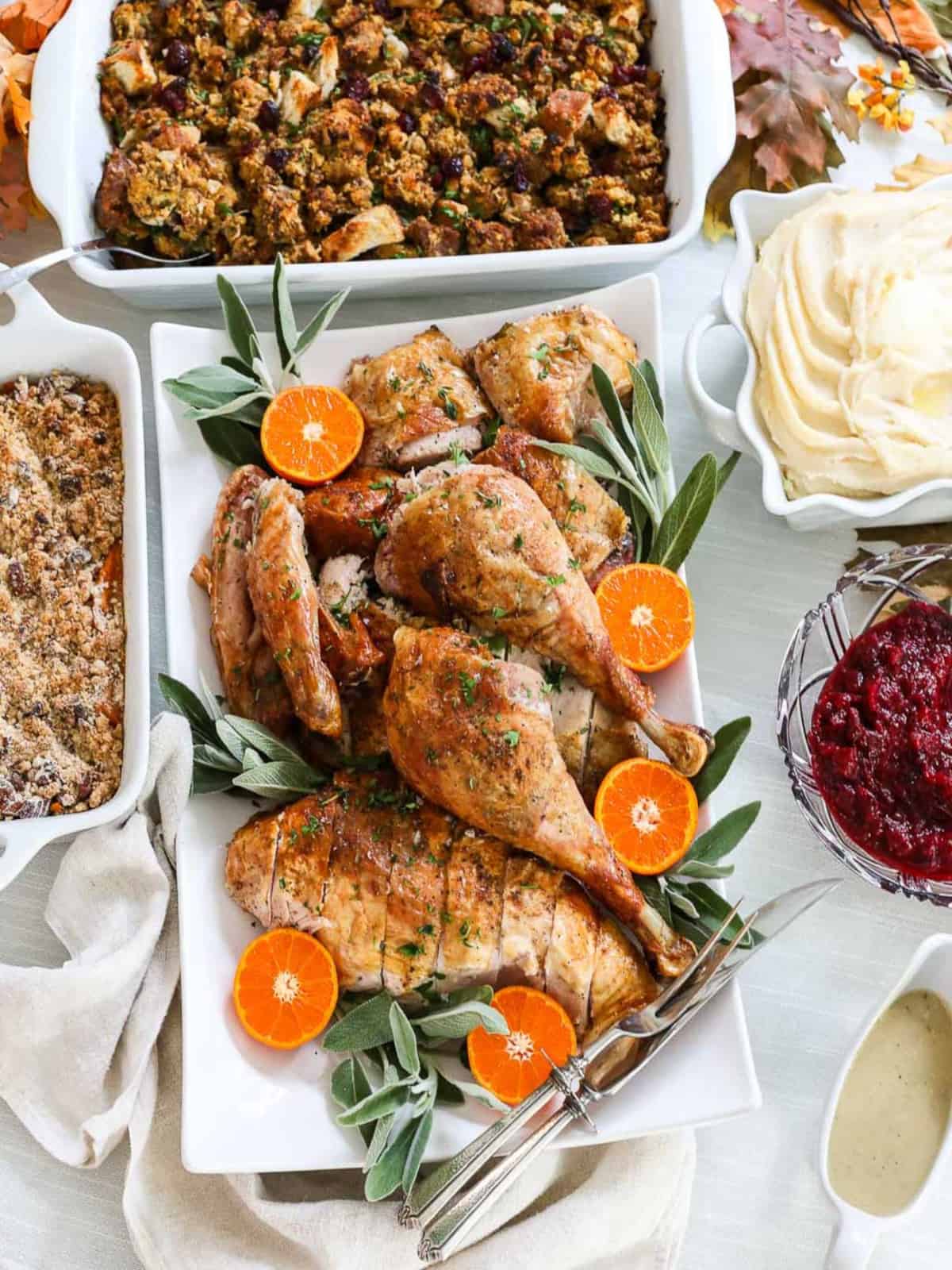 A Thanksgiving dinner table filled with a carved turkey on a platter, sweet potato casserole dish, mashed potatoes, cranberry sauce, gravy, and sausage dressing.