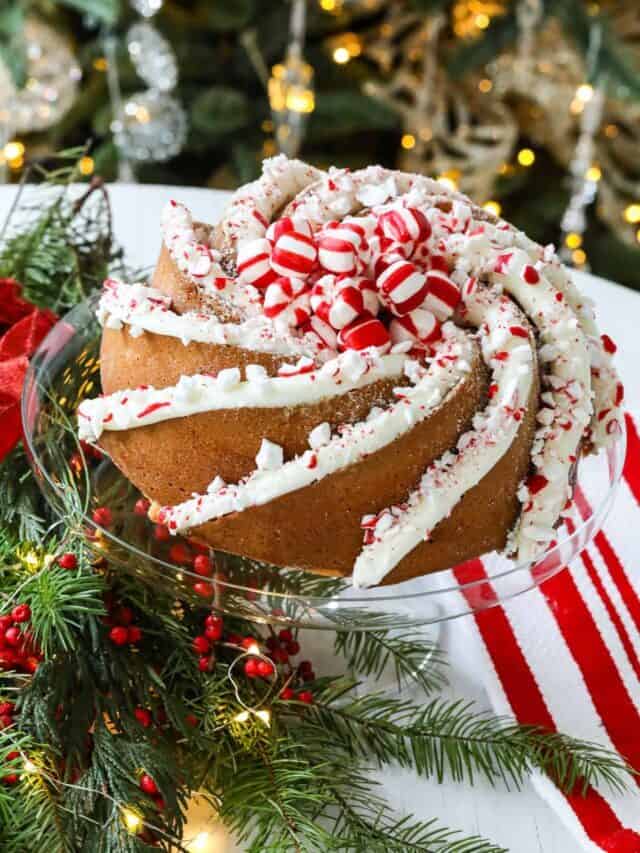 A Christmas bundt cake on a glass cake stand next to holiday greenery with lights and red berries decorated with white buttercream and crushed peppermint candy.