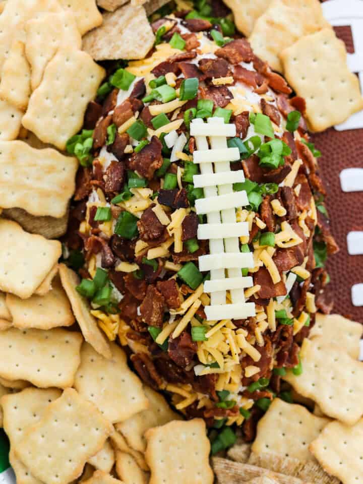 A football shaped cheese ball coated with crumbled bacon, green onions, and cheddar cheese and white cheese to look like laces on the football surrounded by crackers on a platter.