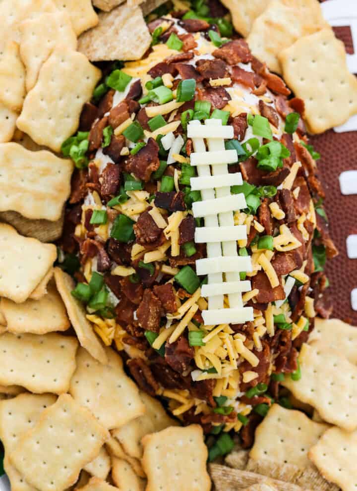 A football shaped cheese ball coated with crumbled bacon, green onions, and cheddar cheese and white cheese to look like laces on the football surrounded by crackers on a platter.