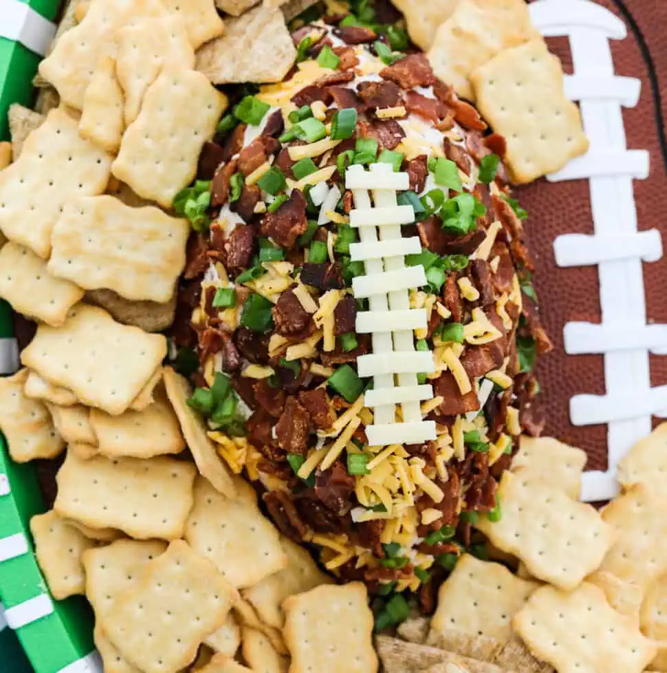A football shaped cheese ball coated with crumbled bacon, green onions, and cheddar cheese and white cheese to look like laces on the football surrounded by crackers on a platter.