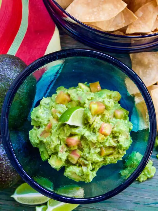 A blue glass bowl filled with guacamole and a chip stuck inside with a bowl of tortilla chips.