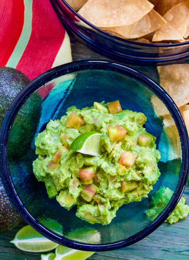 A blue glass bowl filled with guacamole and a chip stuck inside with a bowl of tortilla chips.