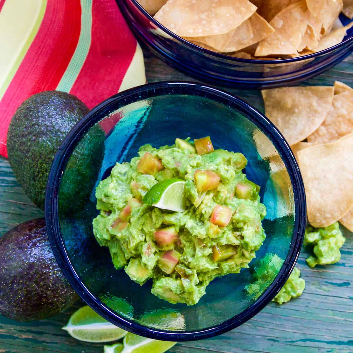 A blue glass bowl filled with guacamole and a chip stuck inside with a bowl of tortilla chips.