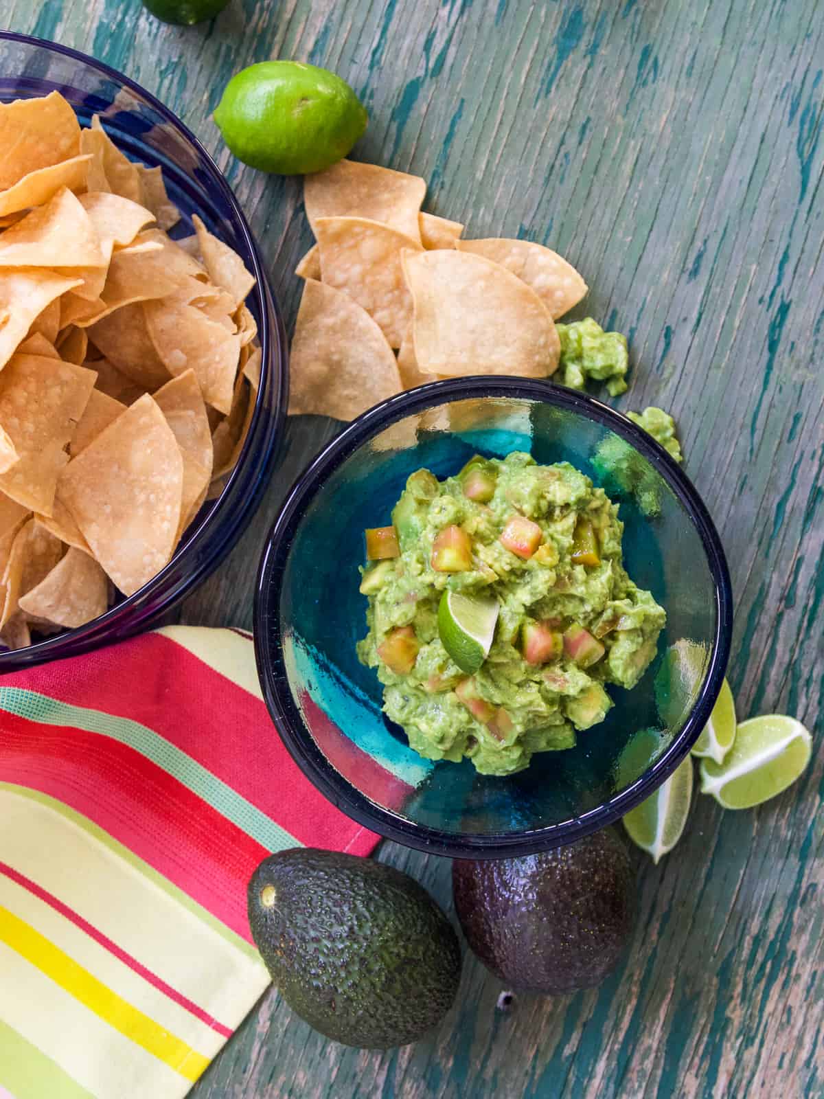 Looking overhead onto a table with a clear blue glass bowl filled with guacamole, and avocados and limes on the table.