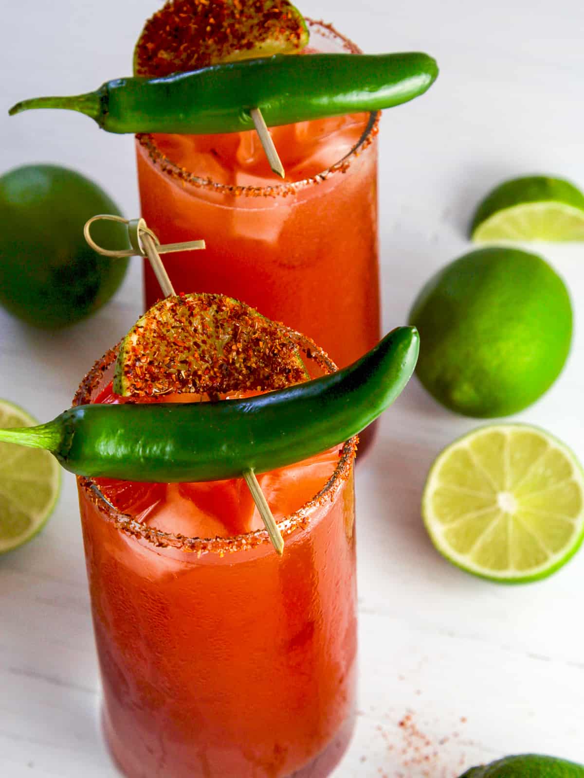 A tall clear glass filled with red Michelada Clamato juice beer cocktail with a green chile pepper garnish on top with seasoned rim on the cocktail.