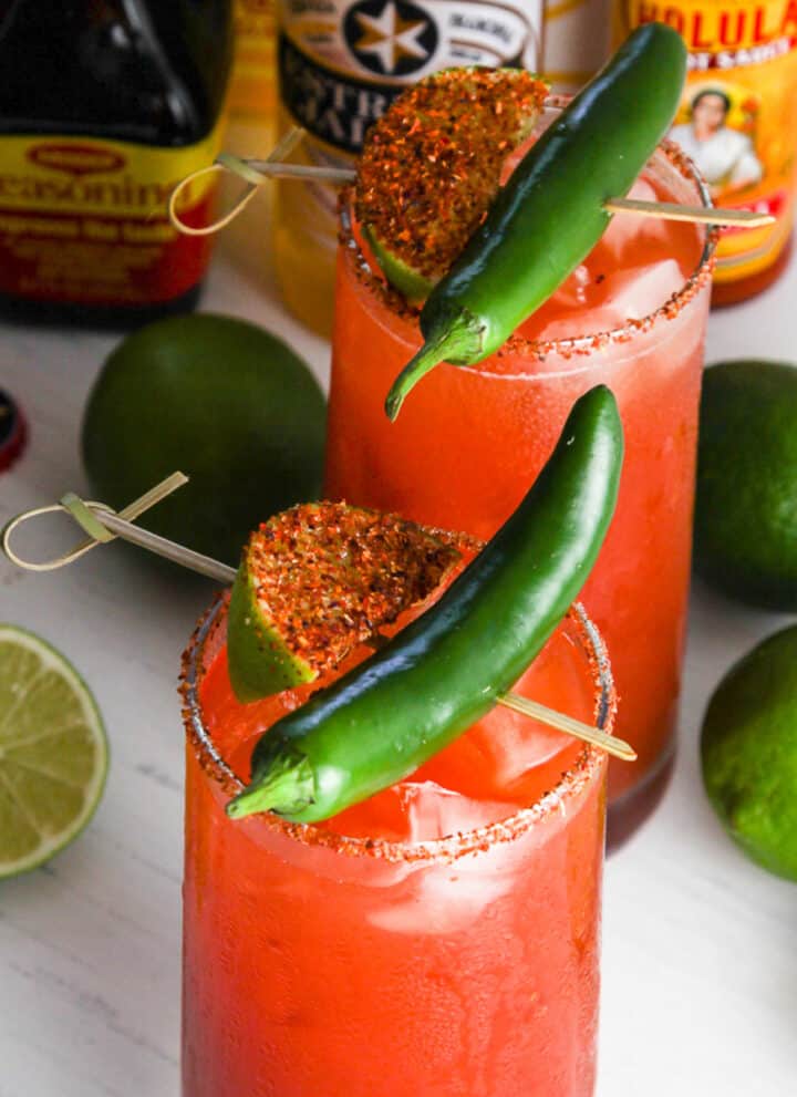 A tall glass filled with michelada garnished with green chili and spice coated lime slice.