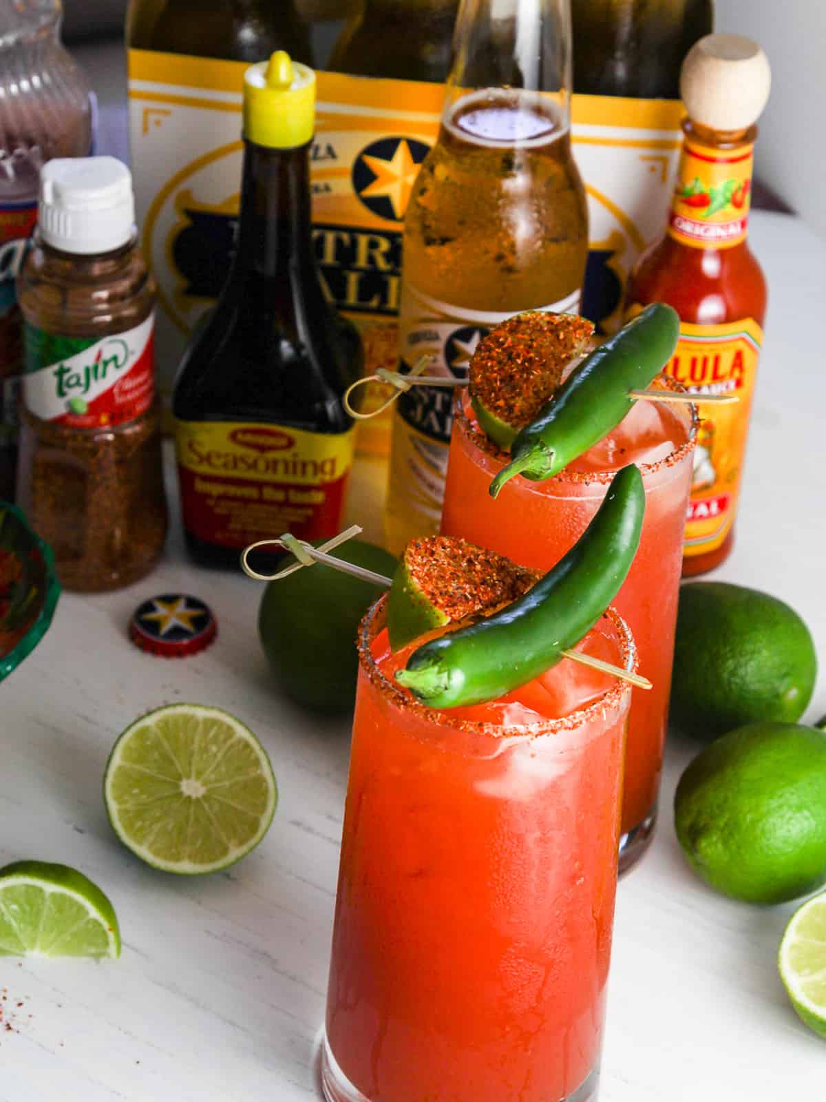 Two tall glasses of Micheladas with a lime sprinkled with Tajin seasoning and berr bottles and limes on the table.