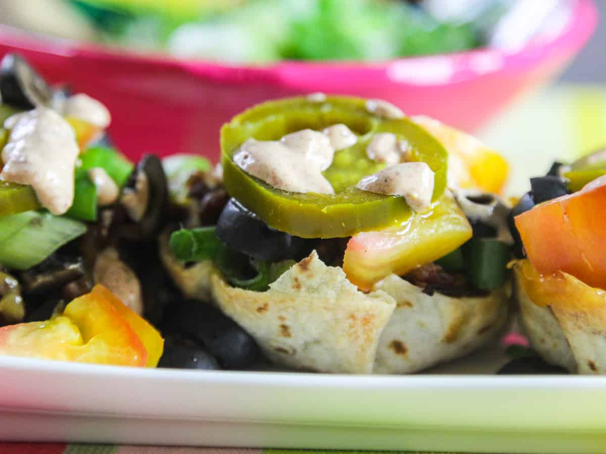Close up of mini tacos in flour tortilla shells topped with cheese, tomato, jalapenos, and black olives.