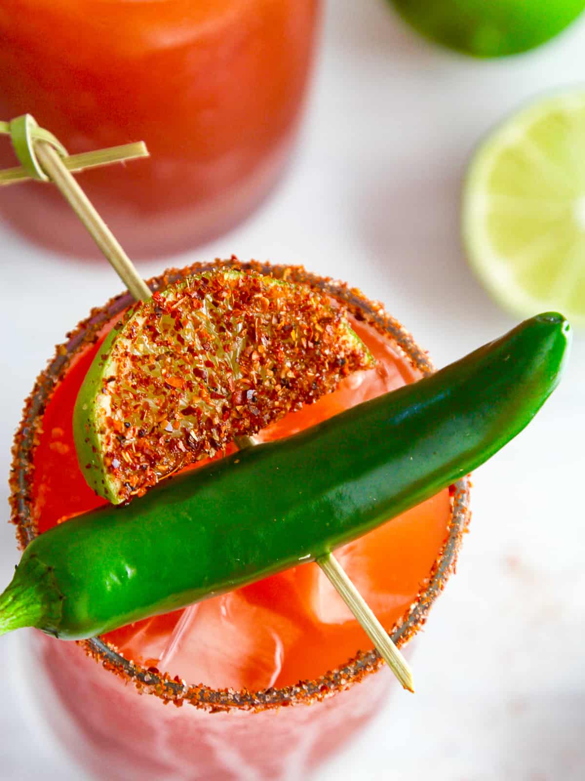 A tall glass filled with michelada garnished with green chili and spice coated lime slice.