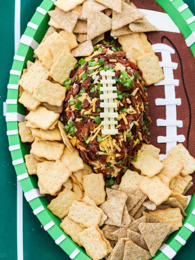 A football shaped cheese ball coated with crumbled bacon, cheddar cheese, and green onions on a plastic tray that looks like a football on a green football field tablecloth.