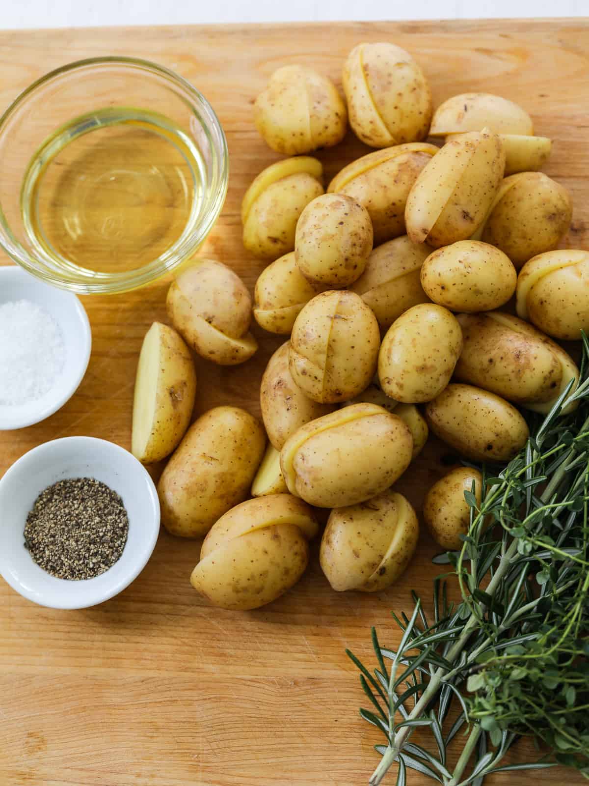 Baby potatoes cut in half on a cutting board with a bunch of fresh herbs, seasoning and olive oil. 