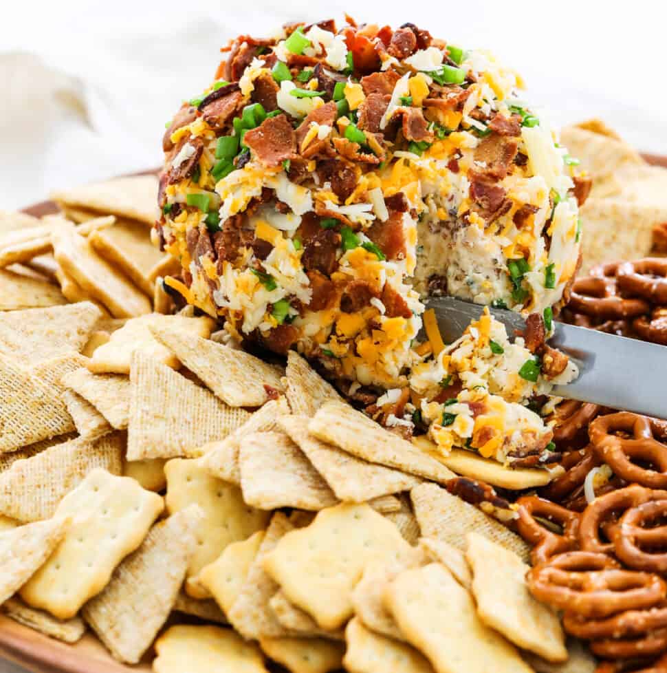 A round bacon cheese ball topped with bacon, cheddar cheese, and surrounded by crackers and pita chips for an easy appetizer.