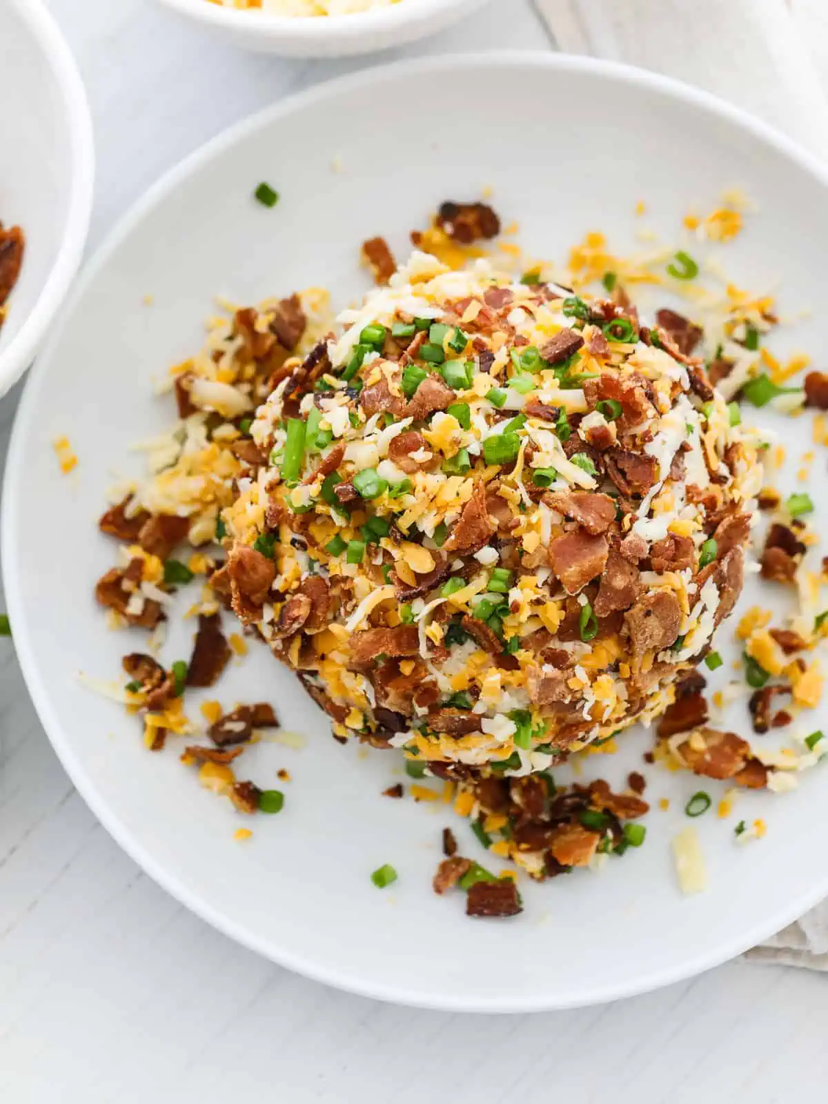 A bacon cheese ball coated on a plate with crumbled bacon, grated cheese, and green onions.