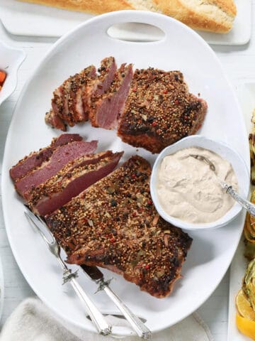 A white platter with sliced corned beef brisket with silver carving set and sauce.