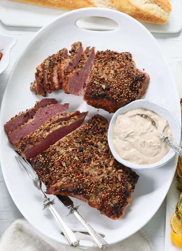 A white platter with sliced corned beef brisket with silver carving set and sauce.