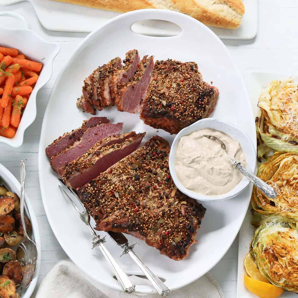 A white platter with sliced corned beef brisket with silver carving set and sauce.