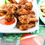 A white platter filled with crispy chicken wings and one dipping into buffalo sauce.
