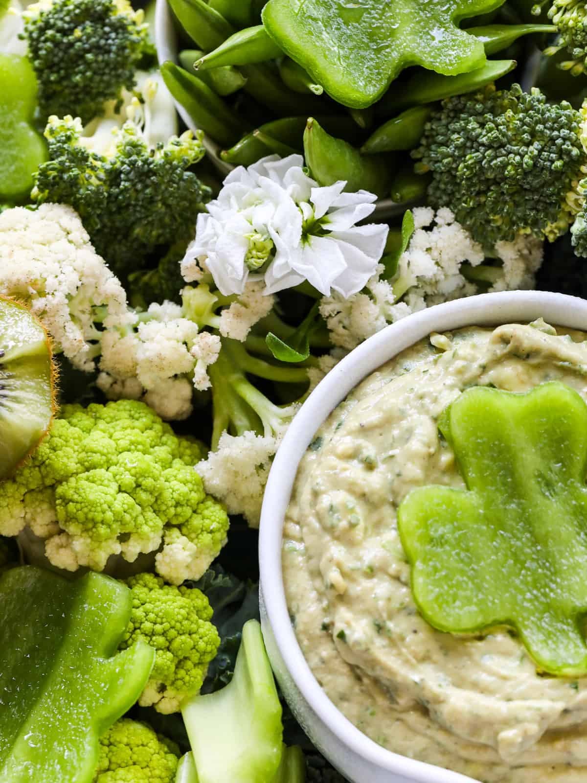 A small white bowl filled with green dip garnished with a clover shape cut out of a green bell pepper with different green fruits and vegetables.