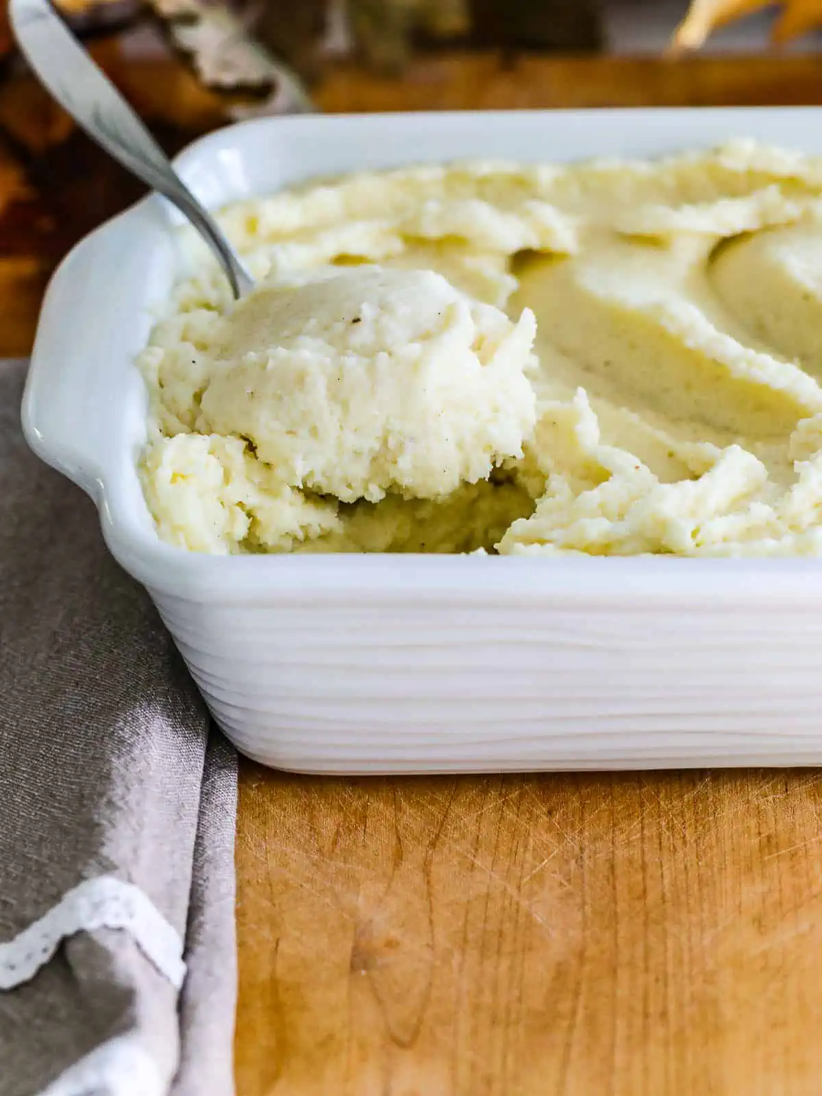 The side of a white casserole dish with a large spoon scooping out a serving of mashed potatoes. 