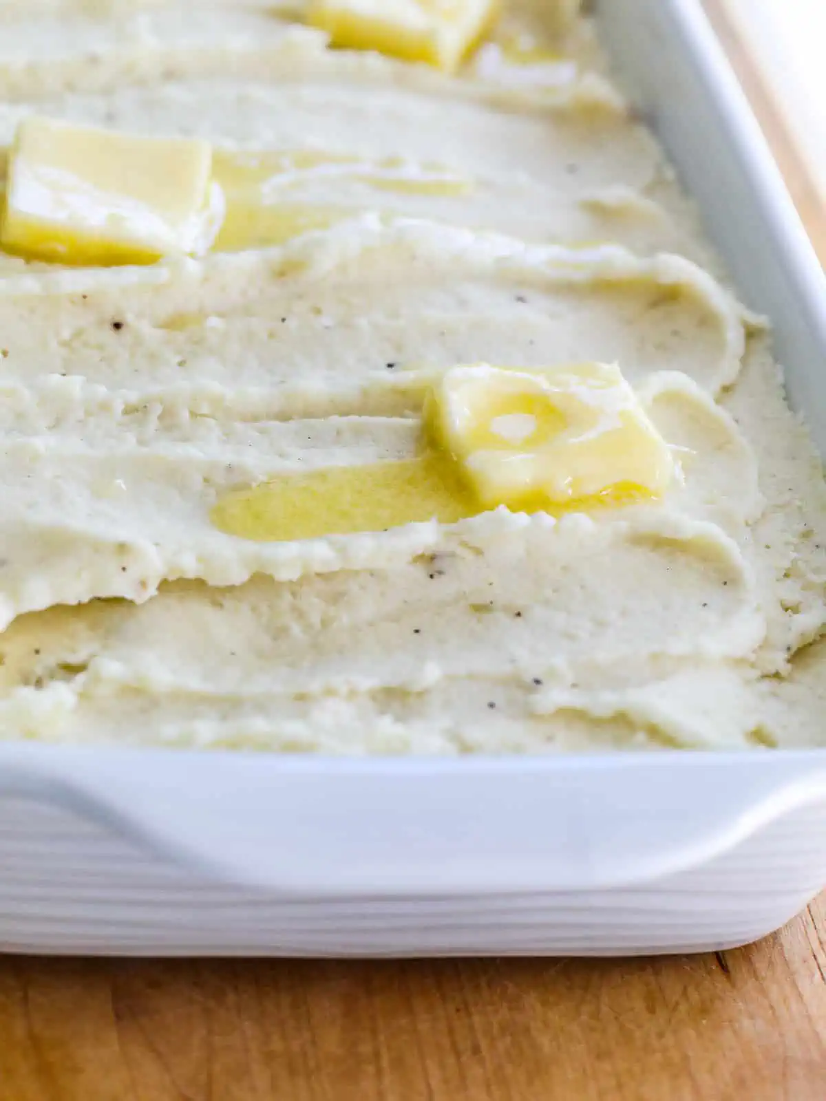 Square pieces of butter melting on hot mashed potatoes in a white casserole dish. 