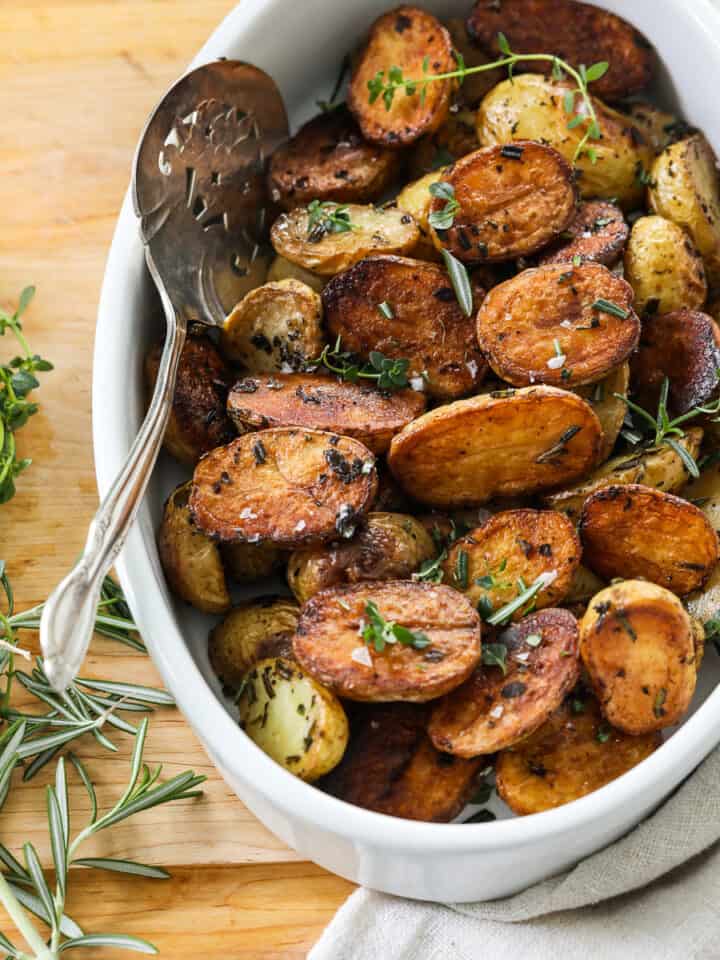A white oval dish filled with golden brown roasted baby potato halves with a silver serving spoon.