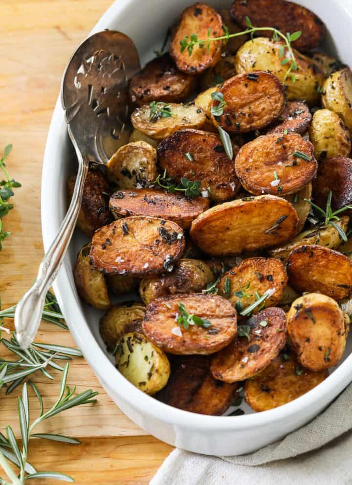 A white oval dish filled with golden brown roasted baby potato halves with a silver serving spoon.