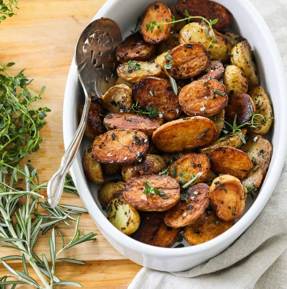 A white oval dish filled with golden brown roasted baby potato halves with a silver serving spoon.