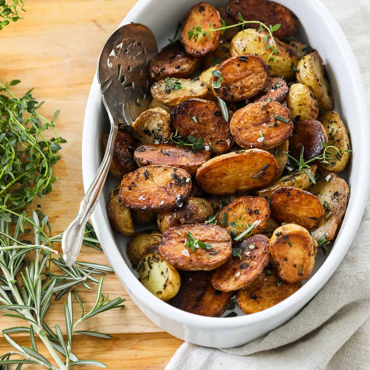 A white oval dish filled with golden brown roasted baby potato halves with a silver serving spoon.