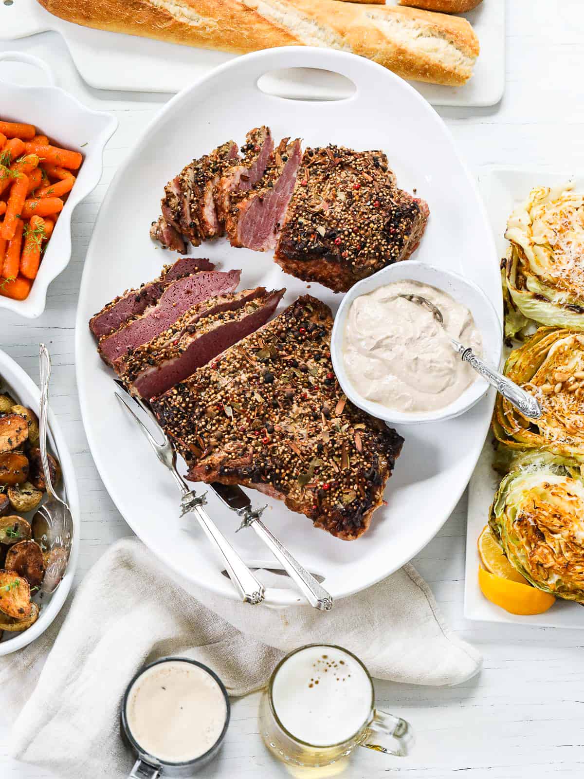 A large white oval platter with corned beef brisket sliced with sauce, beer, and side dishes.