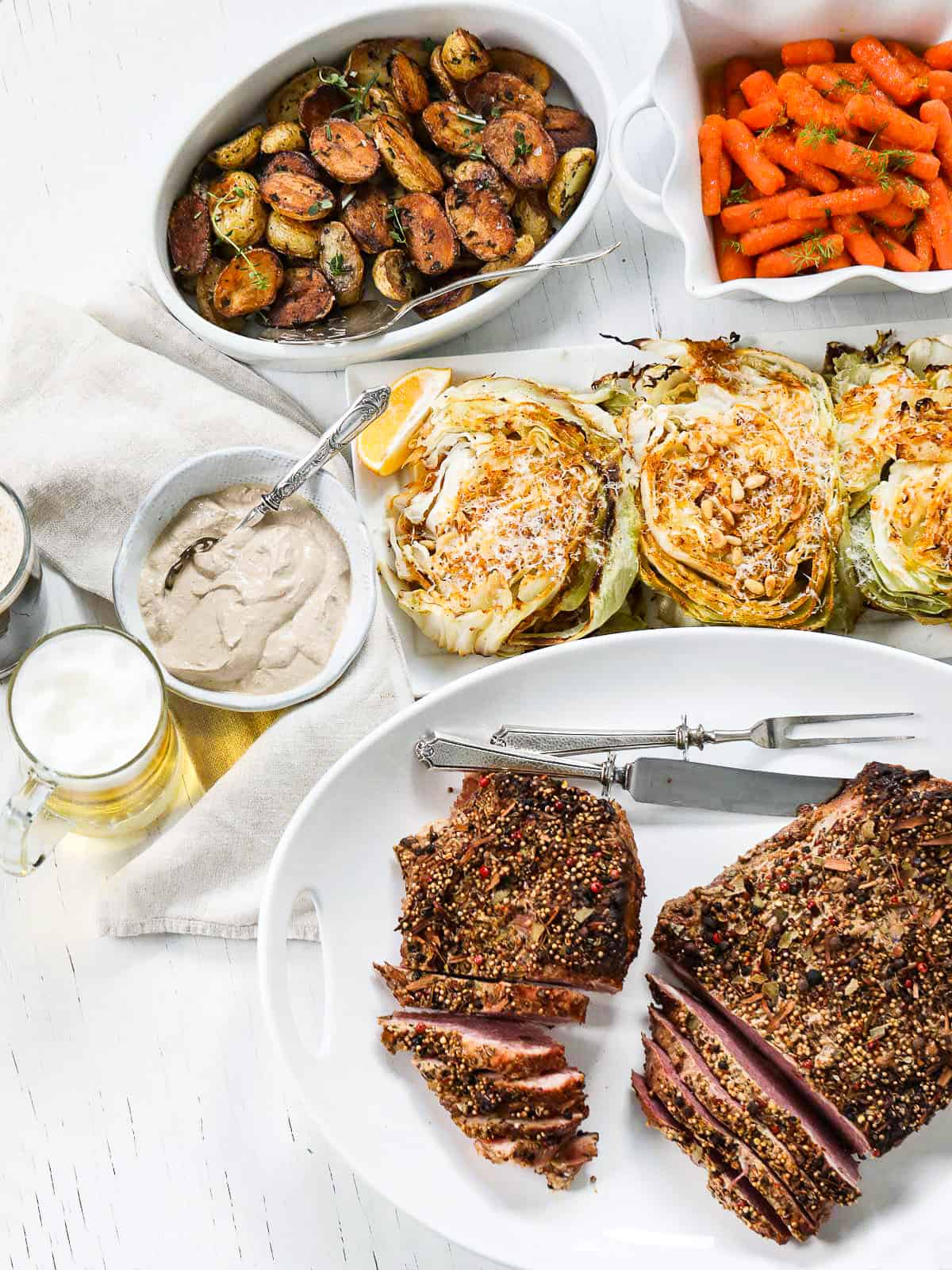 An entire corned beef dinner with sliced beef brisket, roasted cabbage, candied carrots, and roasted potatoes, horseradish cream sauce and glasses of beer. 