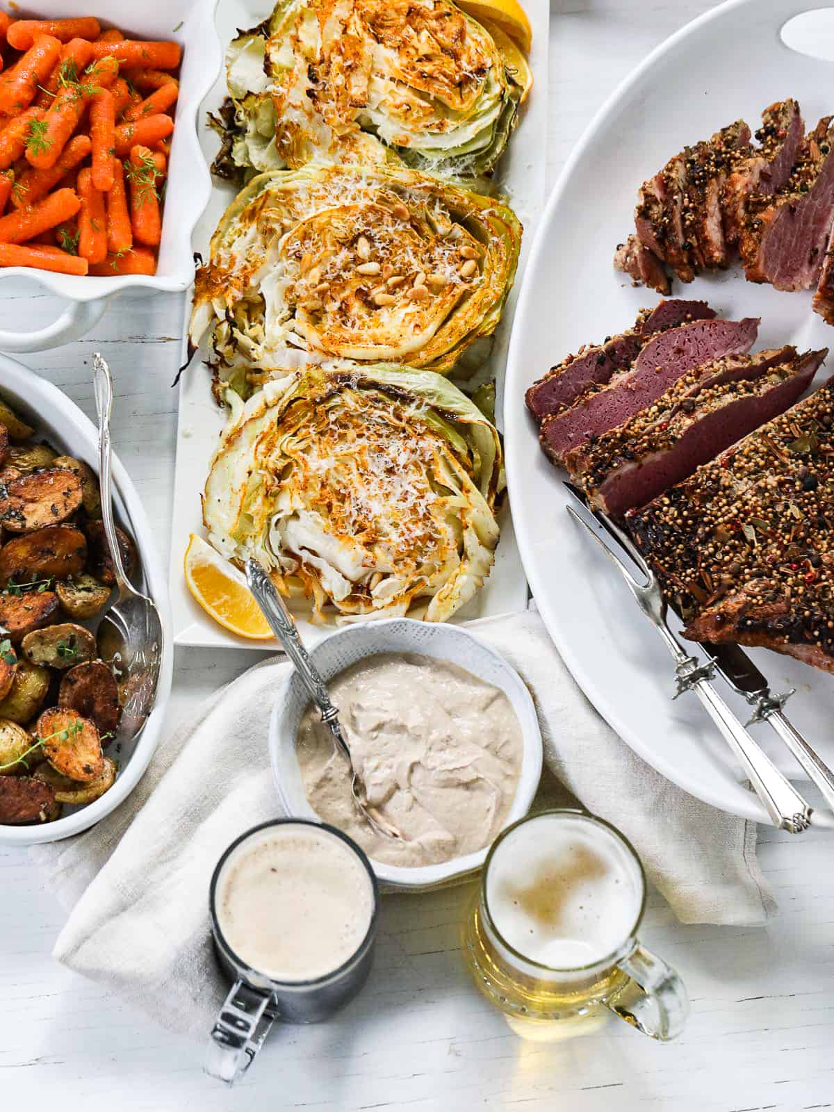 An entire corned beef dinner with sliced corned beef, roasted cabbage, roasted potatoes, glazed carrots, and glasses of beer.