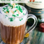 A tall clear coffee mug with a handle with a hot Irish coffee in the mug, topped with white whipped cream, gold sprinkles, and green shamrock sprinkles.