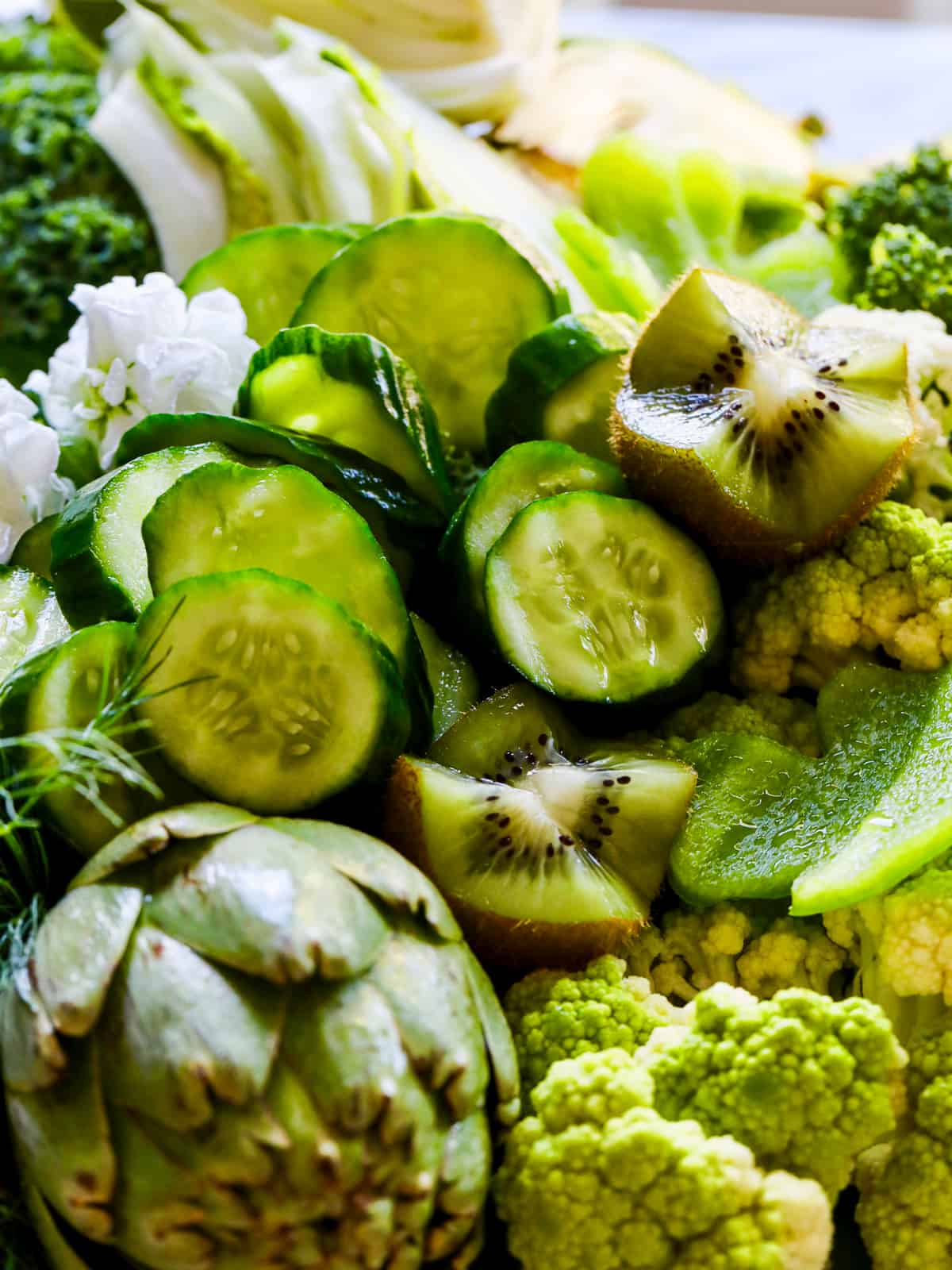 A pile of green vegetables on a tray with slices of cucumber, kiwi, artichokes and more.