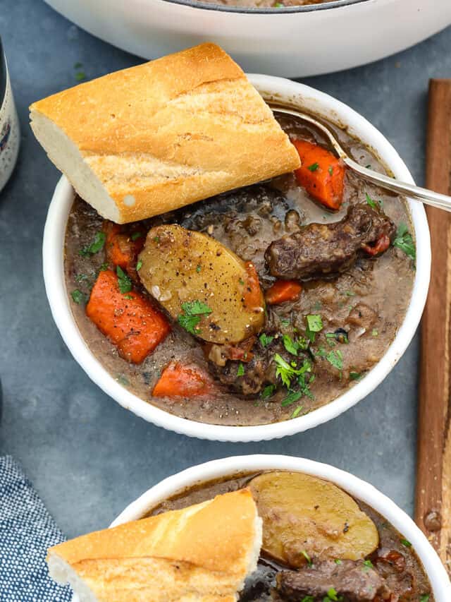 Beef stew in a white bowl with chunks of beef, sliced potatoes, carrots, and slice of baguette with a silver spoon.