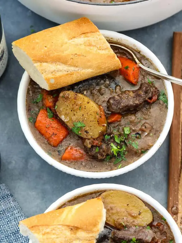 Beef stew in a white bowl with chunks of beef, sliced potatoes, carrots, and slice of baguette with a silver spoon.