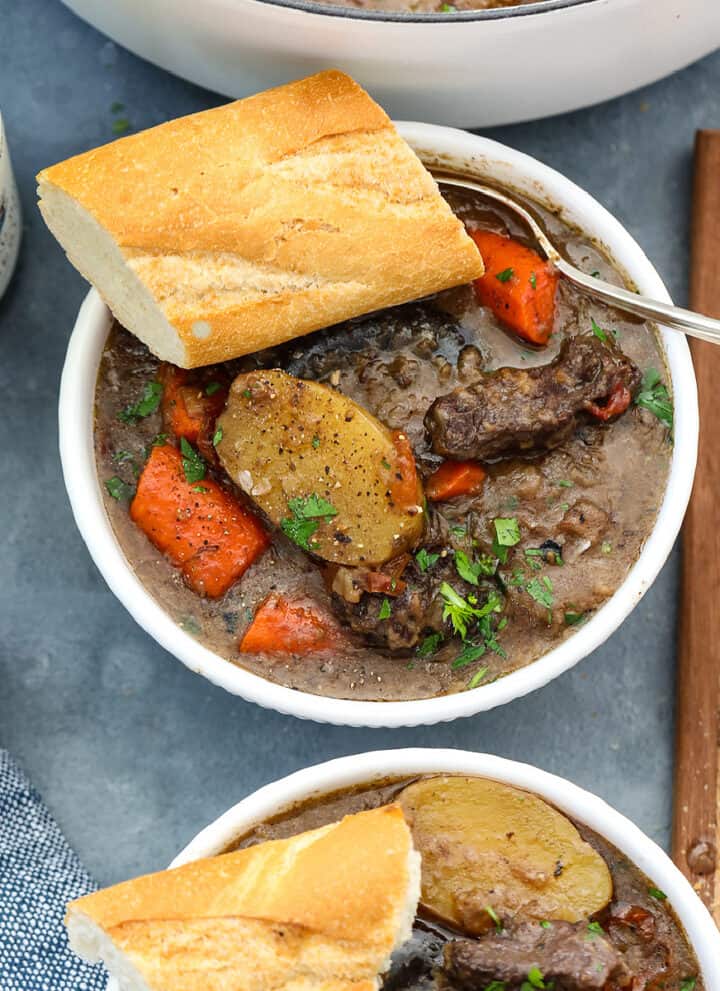 Beef stew in a white bowl with chunks of beef, sliced potatoes, carrots, and slice of baguette with a silver spoon.