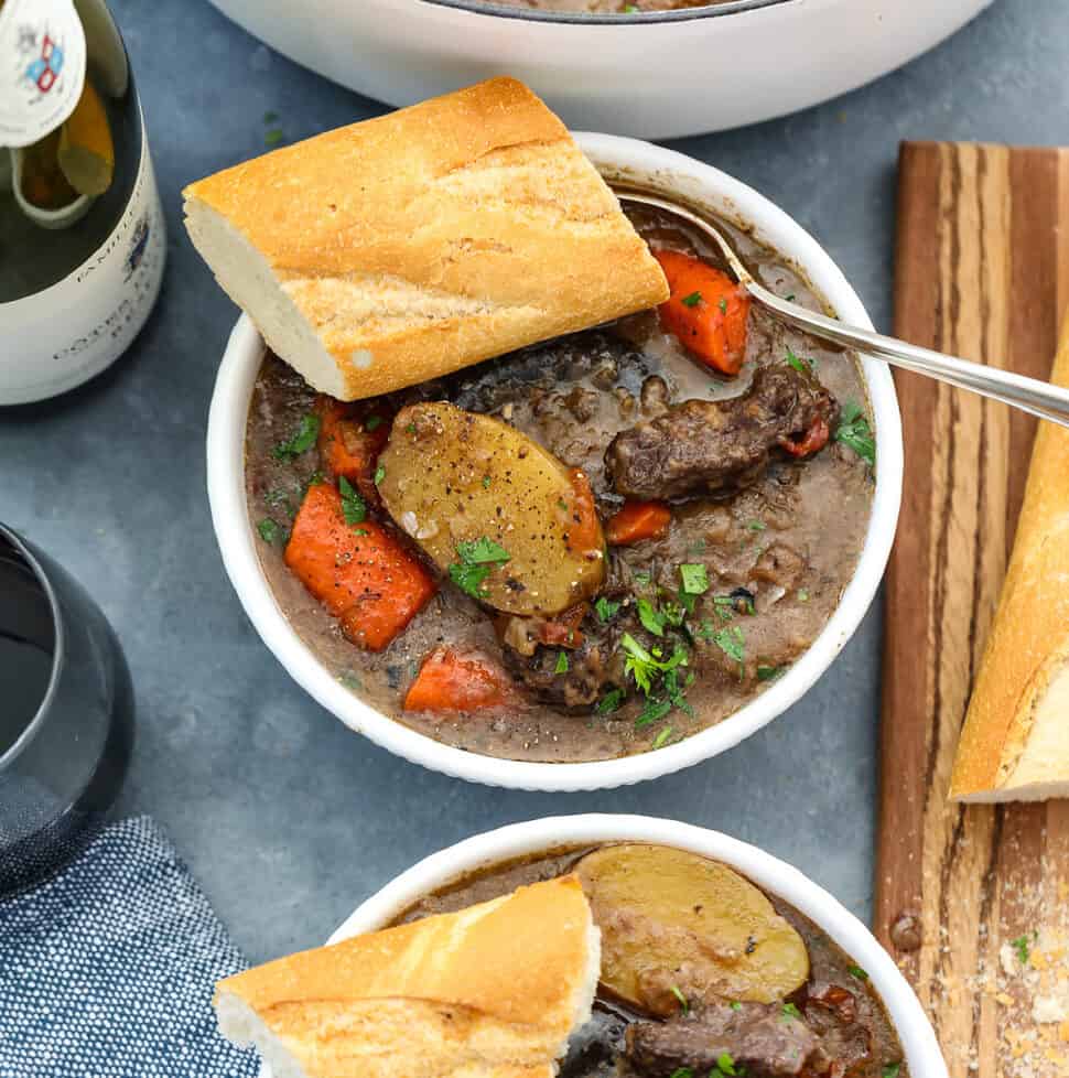 Beef stew in a white bowl with chunks of beef, sliced potatoes, carrots, and slice of baguette with a silver spoon.