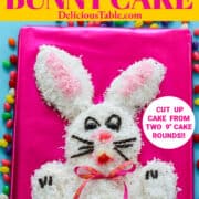 An Easter bunny cake on a hot pink board coated in white coconut, decorated with candy, surrounded by jelly beans.