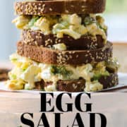 An egg salad sandwich on brown bread stacked on a wood stand with a pickle on top.