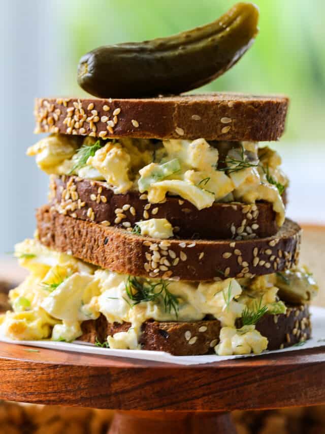 An egg salad sandwich on brown bread stacked on a wood stand with a pickle on top.