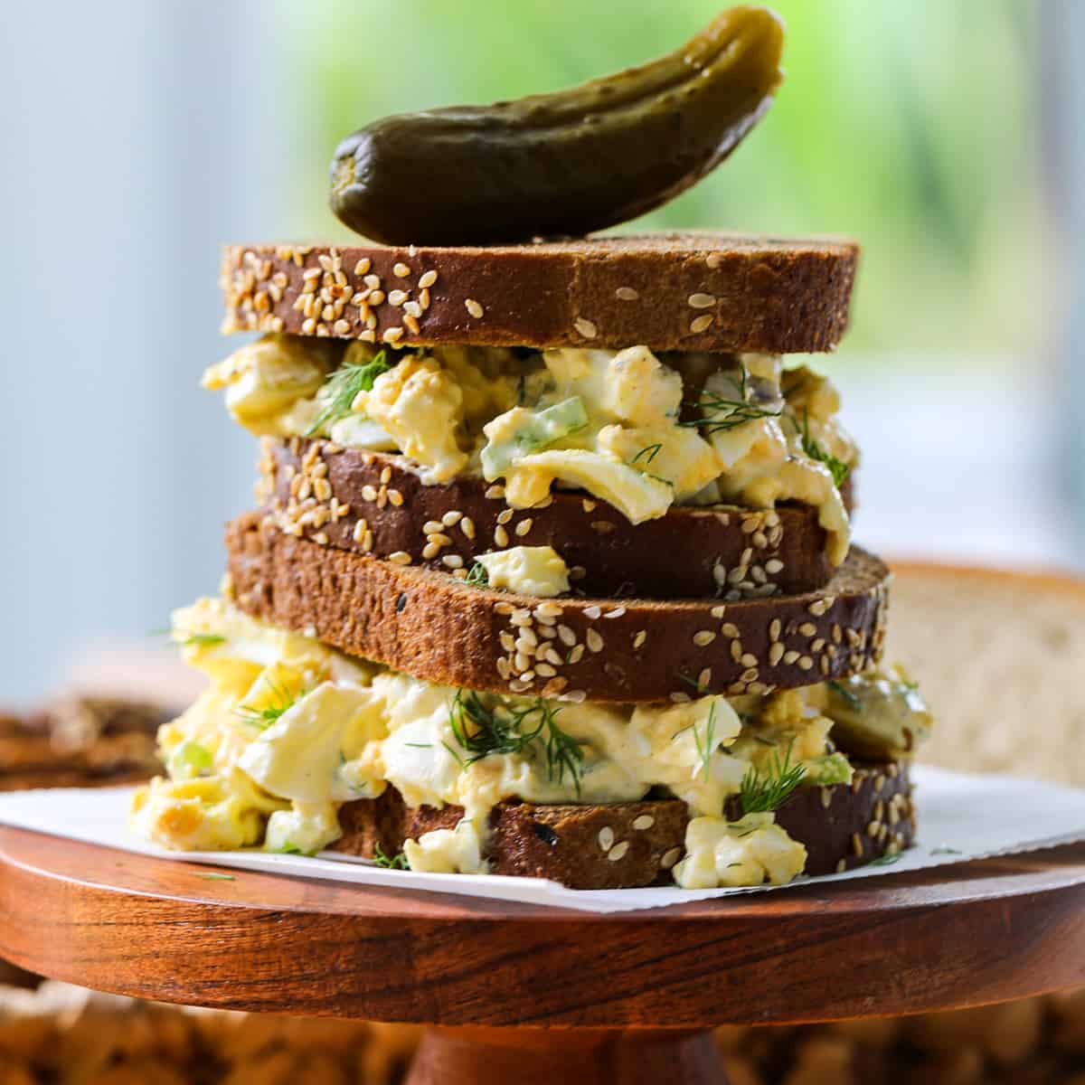 An egg salad sandwich on brown bread stacked on a wood stand with a pickle on top.