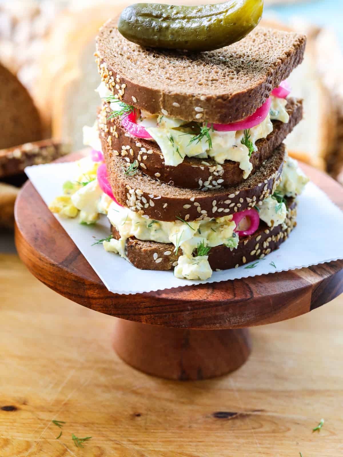 An egg salad sandwich on a wood stand cut in half with a pickle on top and bright pink pickled onions in the sandwich.