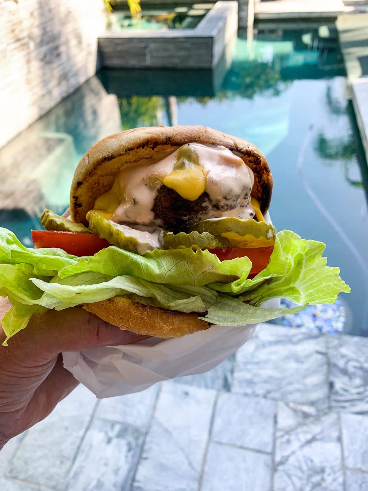 A lady holding a hamburger loaded with burger toppings at a party by the pool.