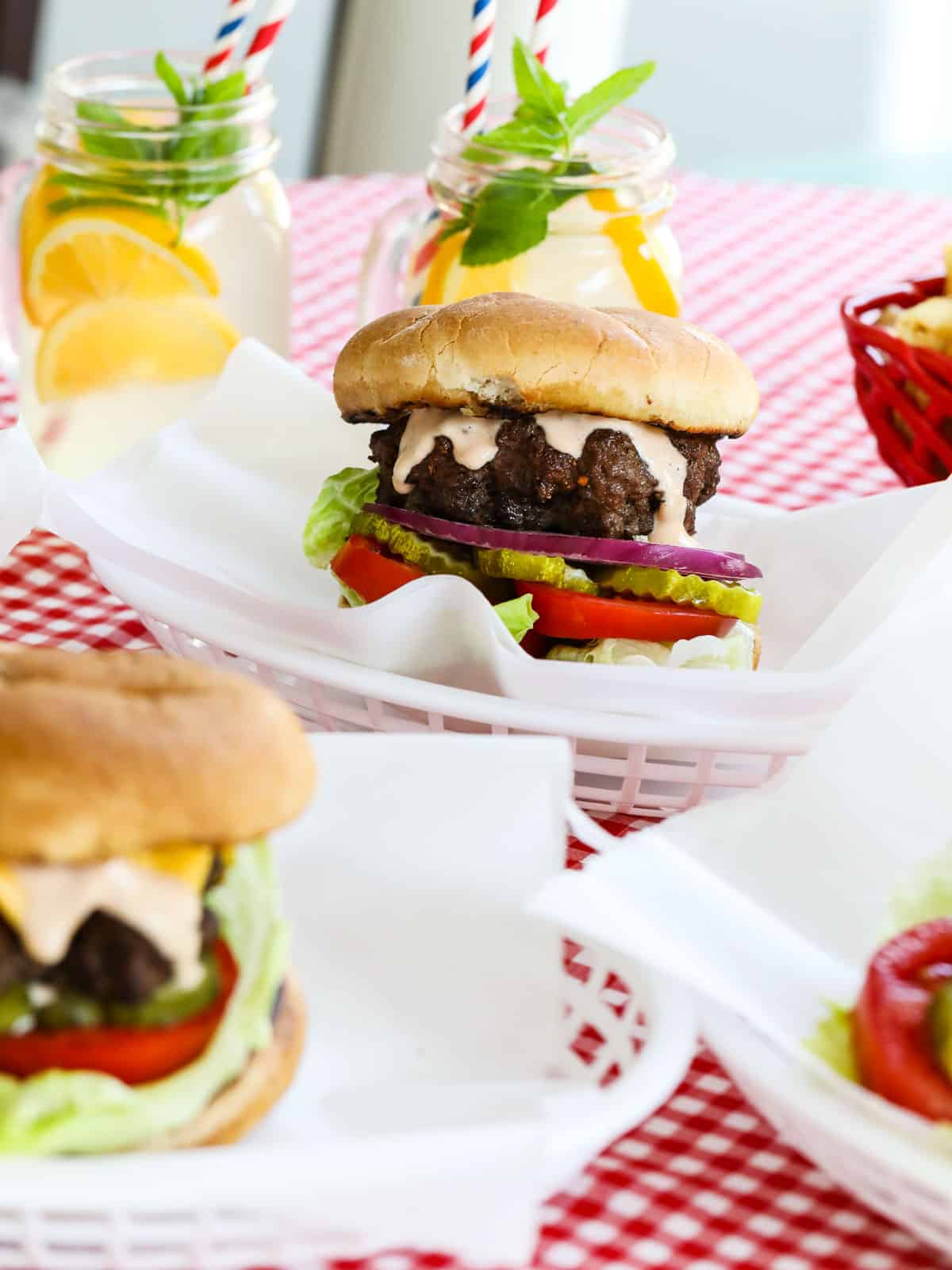 A grilled hamburger recipe in a white food basket with glasses of lemonade. 