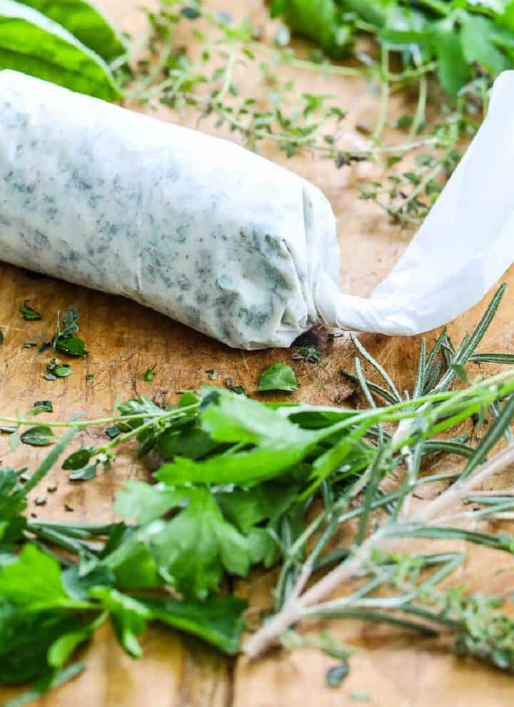 Homemade herb butter wrapped in parchment paper on a cutting board with fresh herbs.