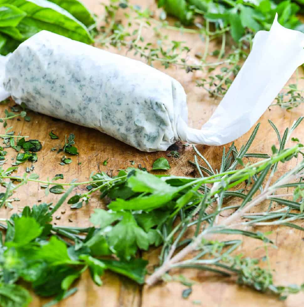 Homemade herb butter wrapped in parchment paper on a cutting board with fresh herbs.