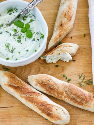 A wooden cutting board with small baguettes sliced open with a white bowl of whipped creamed cheese spread on a slice.