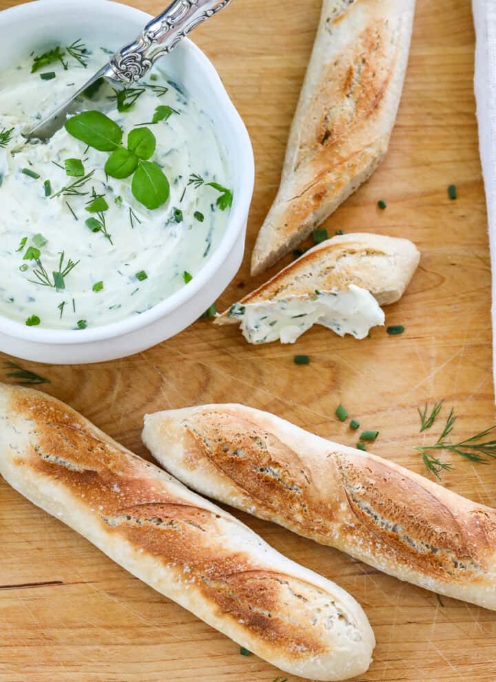 A wooden cutting board with small baguettes sliced open with a white bowl of whipped creamed cheese spread on a slice.